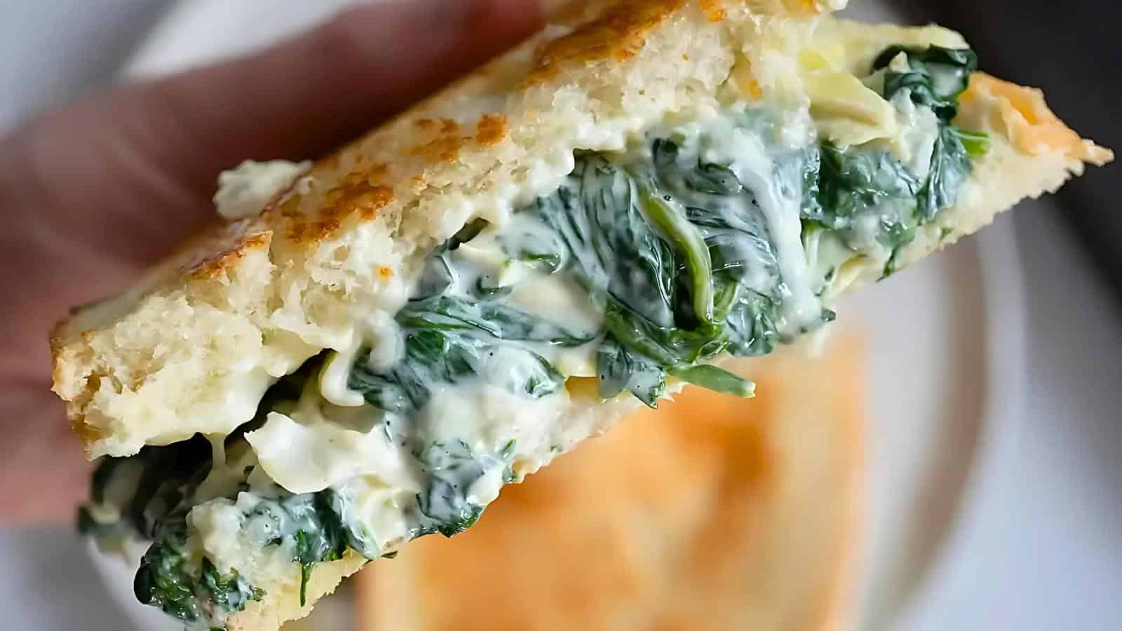 A hand holding a sandwich filled with a creamy spinach and artichoke mixture.