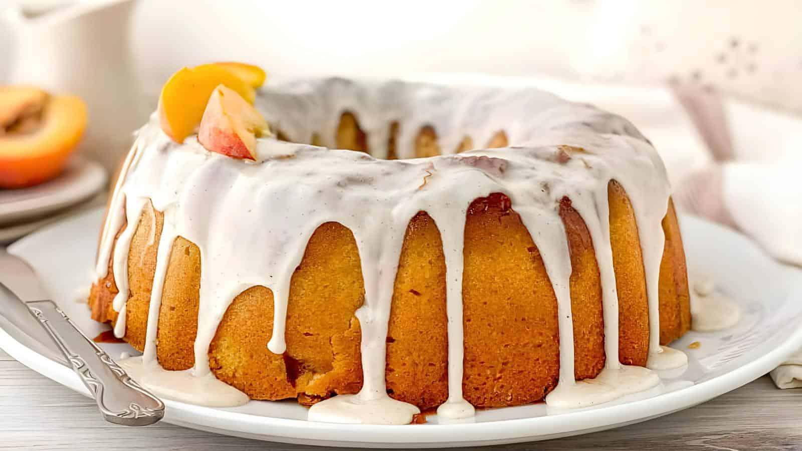 Bundt cake with white icing, garnished with peach slices, on a white plate with a serving knife beside it.