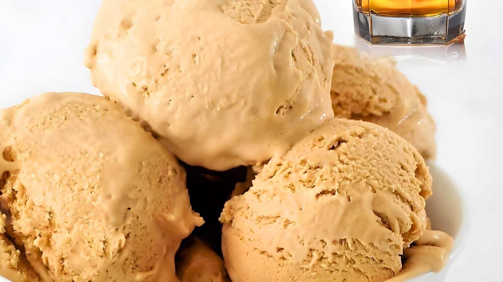 Scoops of caramel-colored ice cream in a white bowl with a glass of whiskey in the background.