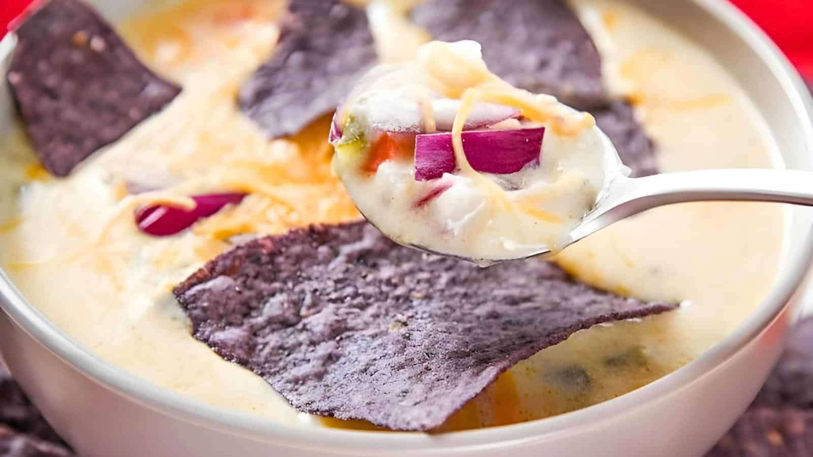 A spoonful of cheesy soup with diced red onion and a purple tortilla chip above a bowl containing more dip and tortilla chips.