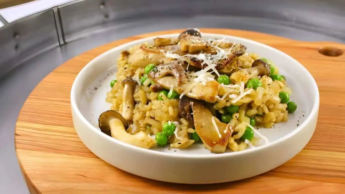 A white plate filled with creamy mushroom risotto garnished with green peas and shaved Parmesan cheese. The dish is presented on a wooden serving board. The background is blurred, emphasizing the colorful and appetizing risotto in the foreground.