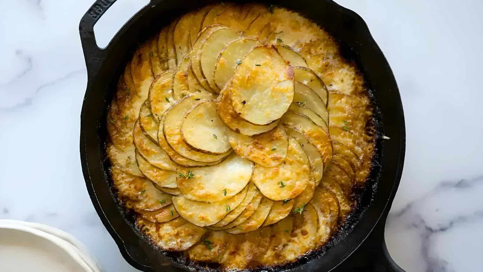 A cast iron skillet filled with golden-brown scalloped potatoes, topped with fresh herbs.