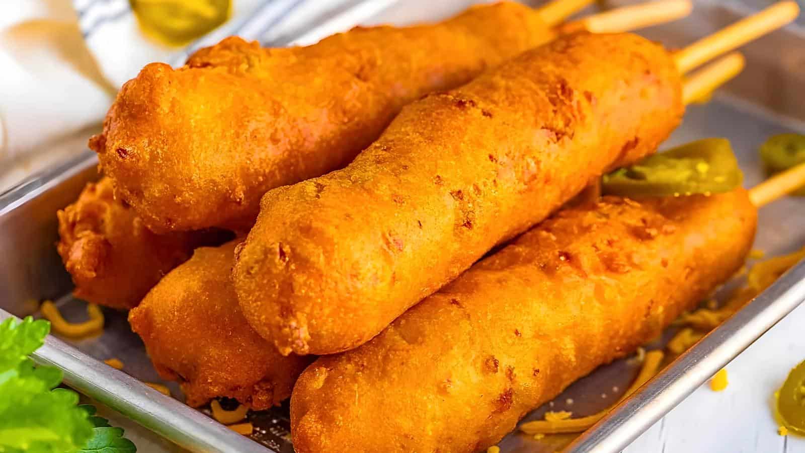 Five golden-brown corn dogs on wooden sticks are placed on a rectangular metal tray, garnished with sliced jalapeños.