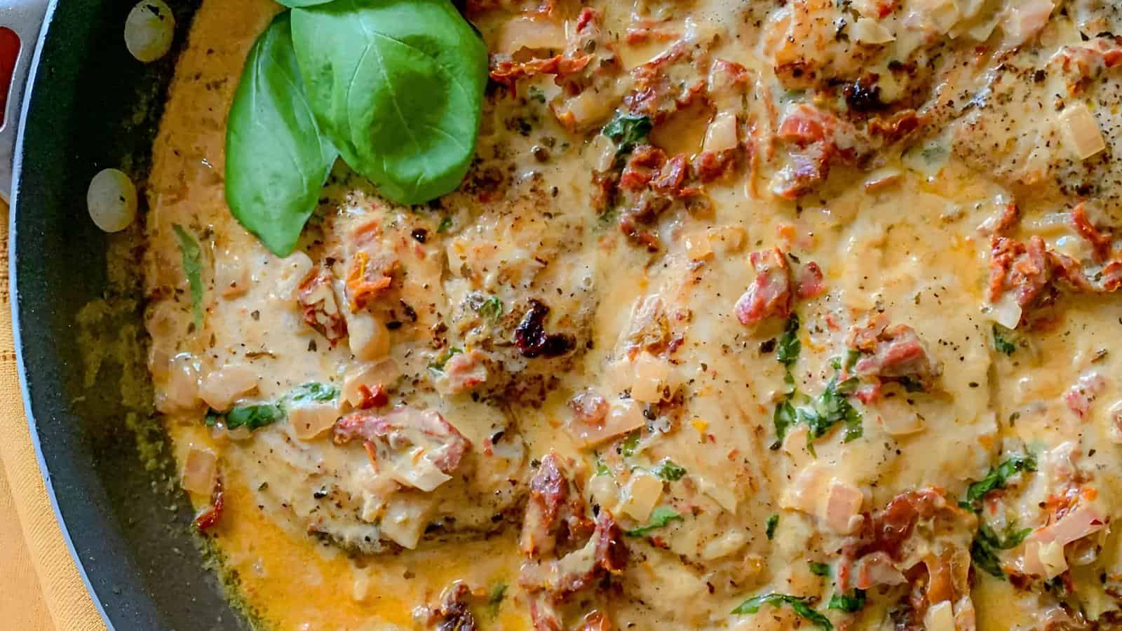 A skillet filled with creamy chicken, garnished with fresh basil leaves, featuring sun-dried tomatoes, a creamy sauce, chunks of chicken, and herbs.