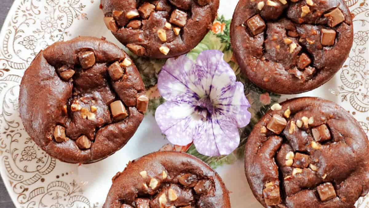 A decorative plate holds six chocolate muffins topped with small chocolate chunks and nuts. In the center of the plate, there is a purple flower, adding a touch of elegance to the arrangement. The plate has intricate, delicate patterns around the edge.