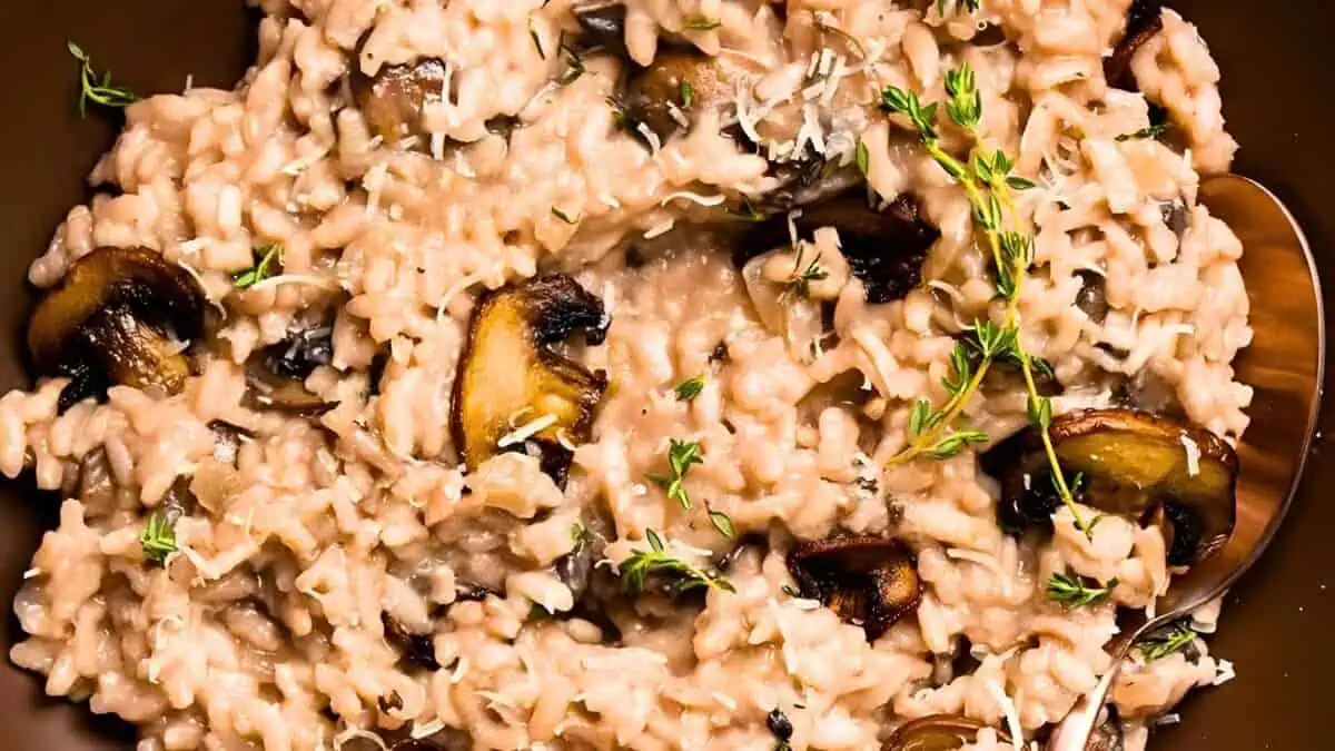 A close-up of a creamy mushroom risotto garnished with fresh thyme. The risotto features plump grains of rice, tender mushroom slices, and is topped with a sprinkling of grated cheese. The dish is displayed in a bowl with a spoon on the side.