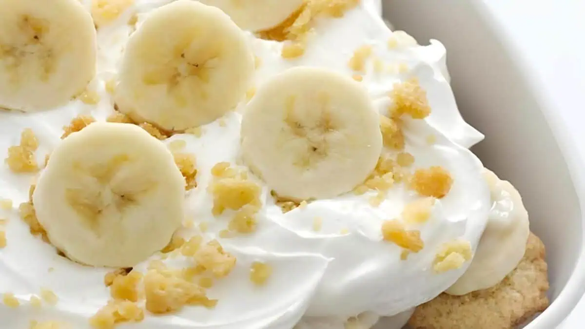 A close-up shot of a dessert featuring slices of banana and crumbled cookies on top of a layer of creamy white whipped topping. The dessert is presented in a white bowl.