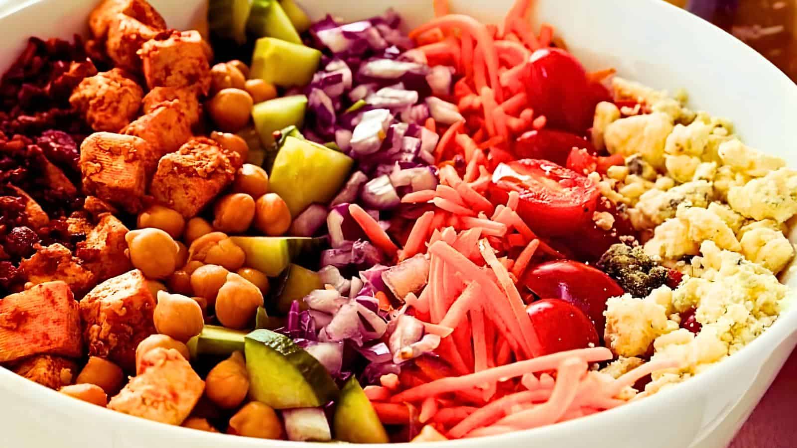 A white bowl filled with rows of chickpeas, diced cucumbers, red cabbage, grated carrots, cherry tomatoes, and crumbled feta cheese.