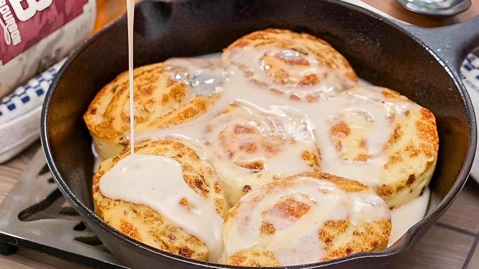 A cast iron skillet filled with cinnamon rolls being topped with icing, with a bag of flour in the background.