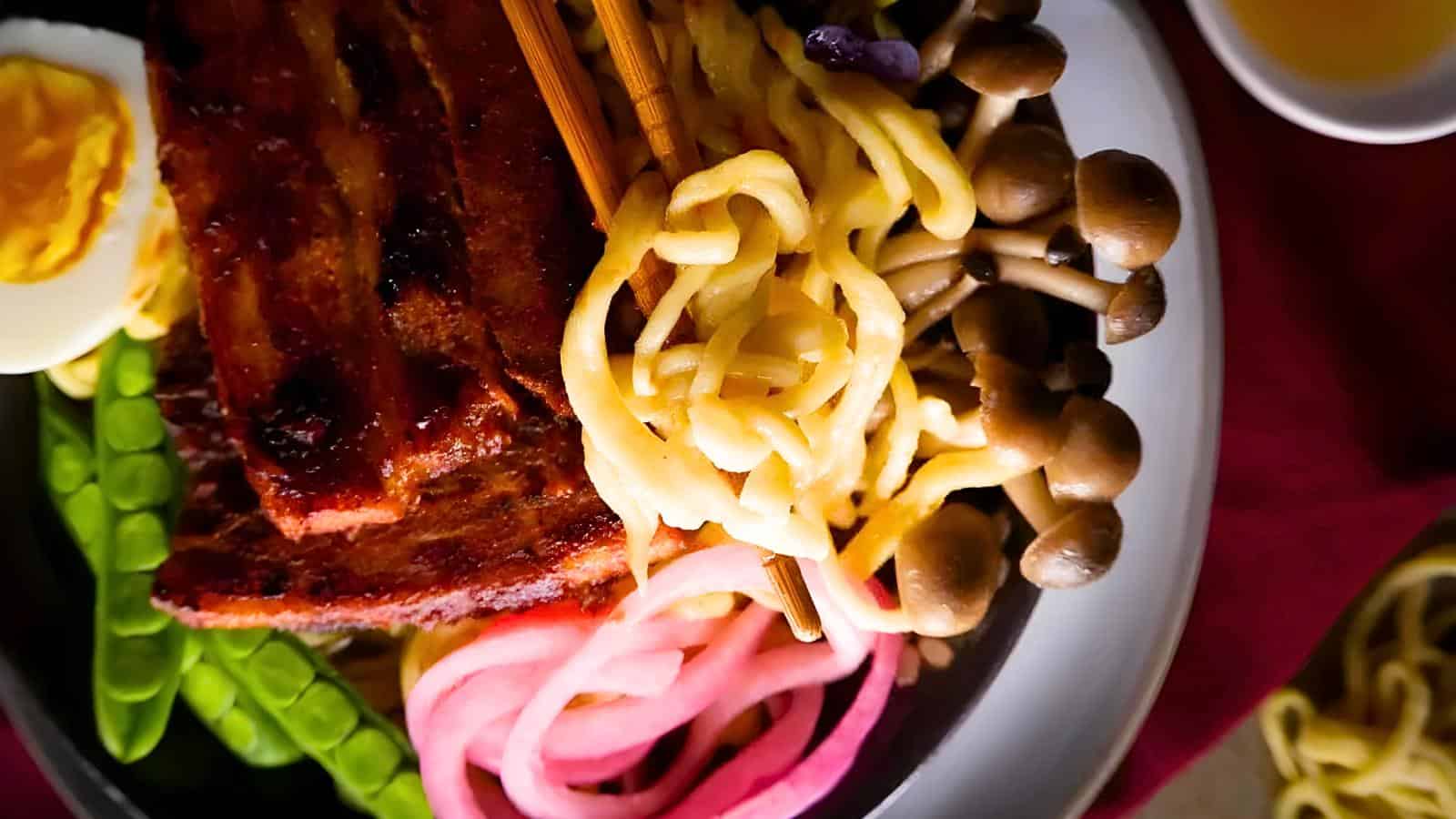 A bowl of ramen topped with sliced pork, a halved egg, mushrooms, pickled onions, and snap peas. Chopsticks rest on top of the noodles.