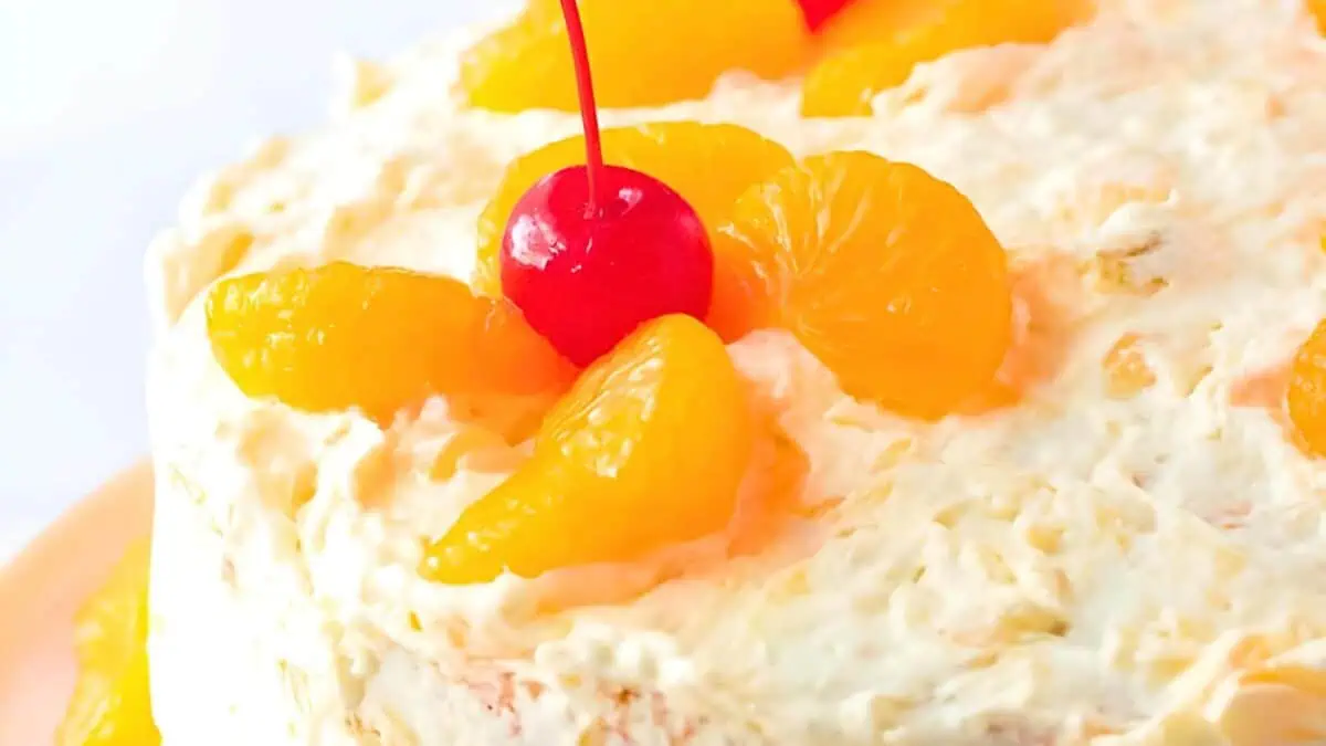 A close-up view of a cake topped with creamy frosting and garnished with bright orange mandarin slices and a vivid red cherry in the center. The cake appears to have a light, fluffy texture with a citrusy, refreshing look.