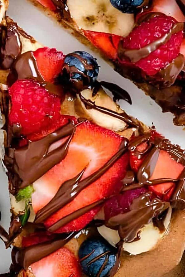A close-up of pizza topped with strawberries, blueberries, banana slices, and raspberries, drizzled with chocolate sauce.