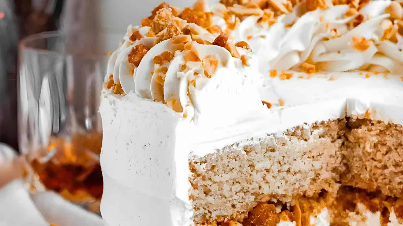 A close-up of a cake with cream-colored frosting topped with crumbled pieces and a slice missing, displaying its dense, moist texture. The blurred background shows glassware.