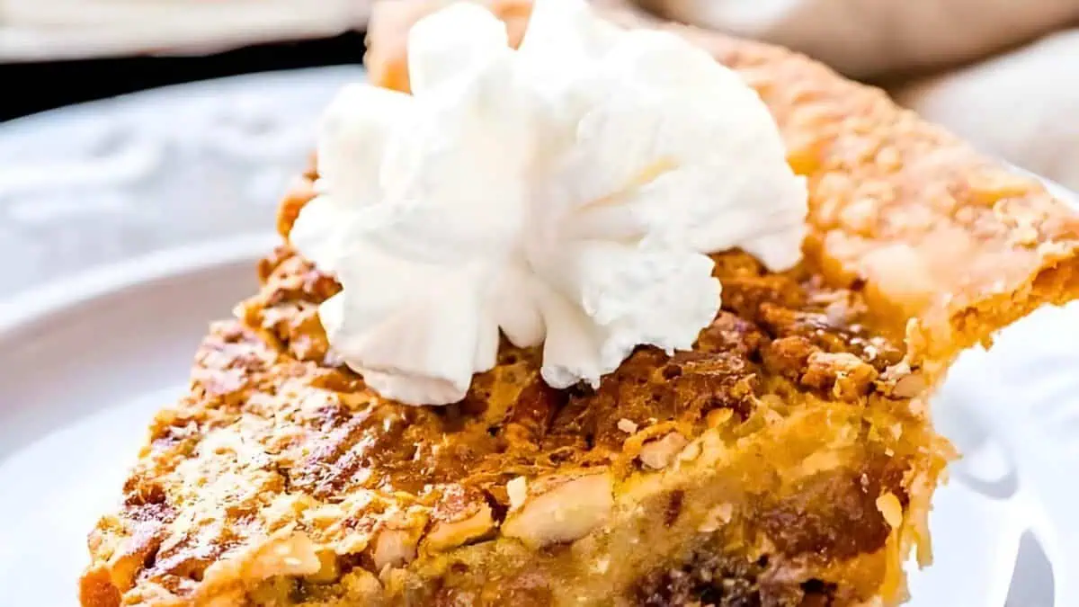 A close-up of a slice of pecan pie topped with a dollop of whipped cream on a white plate. The pie's filling appears rich and gooey, with a crispy, golden crust.