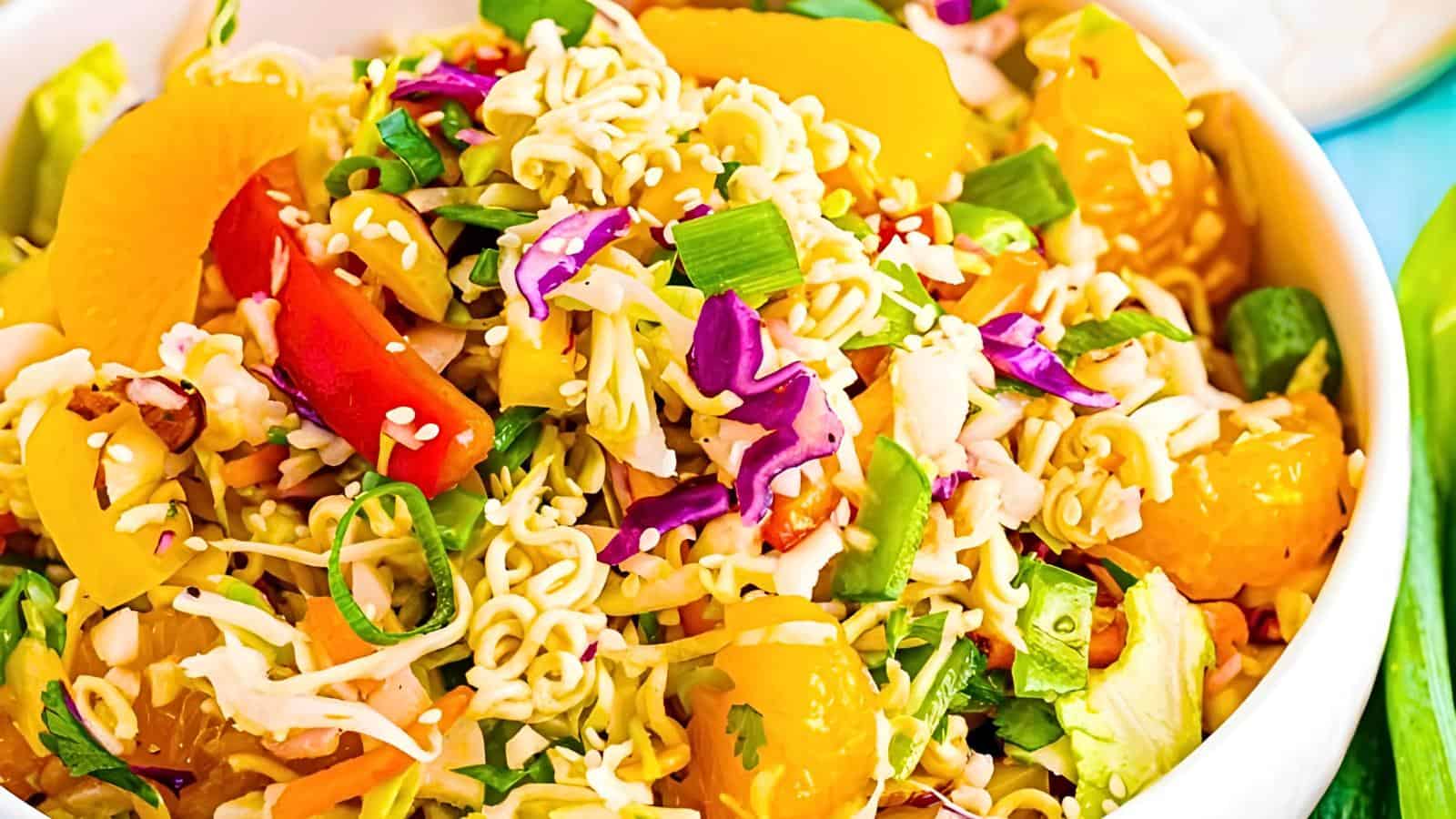 A vibrant bowl of salad featuring colorful ingredients such as orange mandarin slices, red and purple cabbage, green onions, noodles, and bell peppers, topped with sesame seeds and a light dressing.
