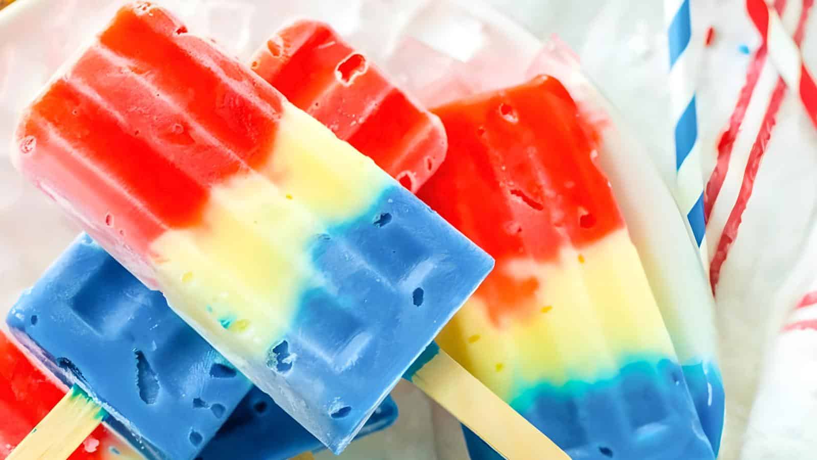 Colorful popsicles with red, yellow, and blue layers on sticks, placed on a light background with striped straws.