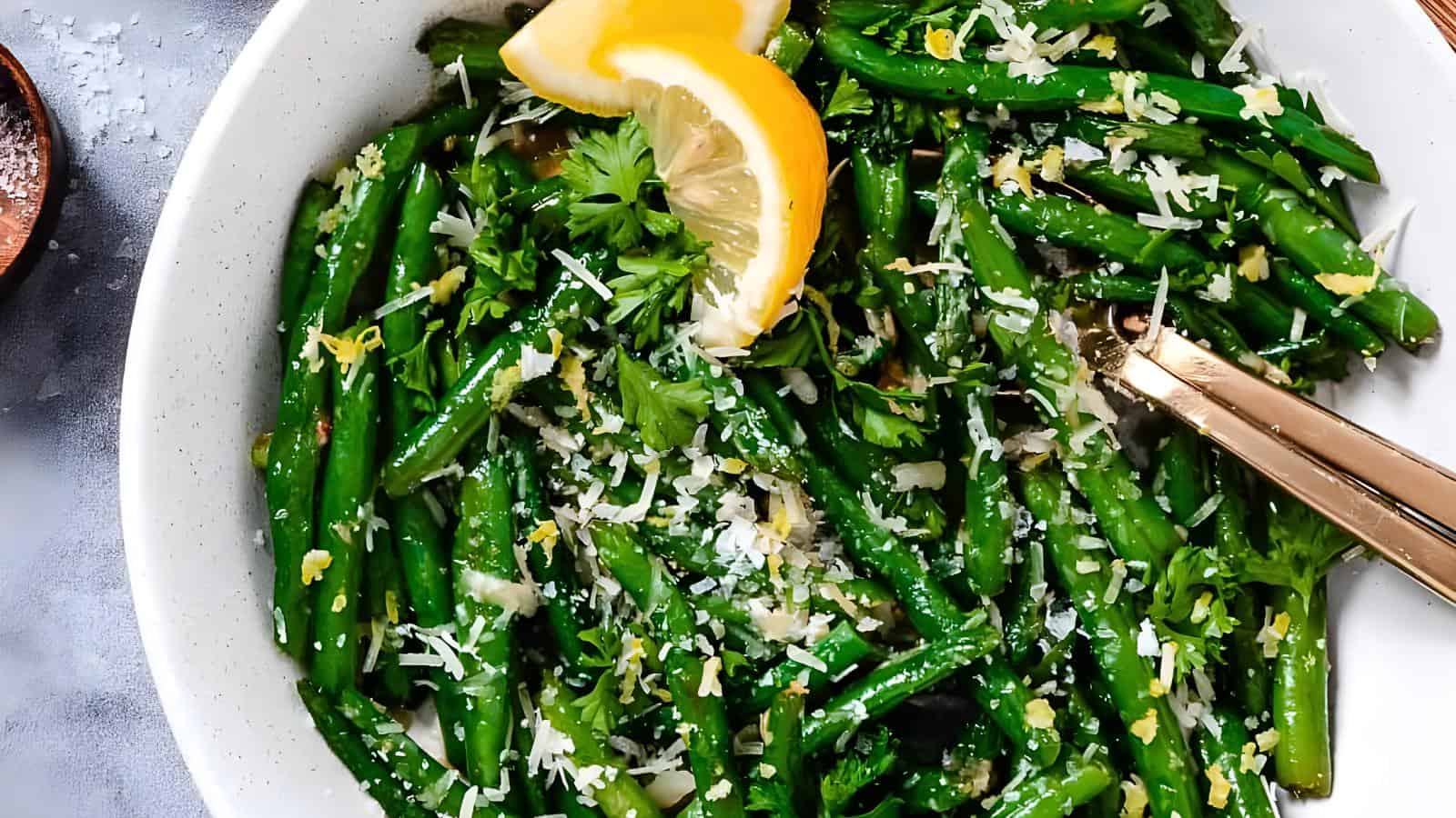 A bowl of cooked green beans garnished with grated cheese, parsley, and lemon slices, with gold-colored utensils on the side.