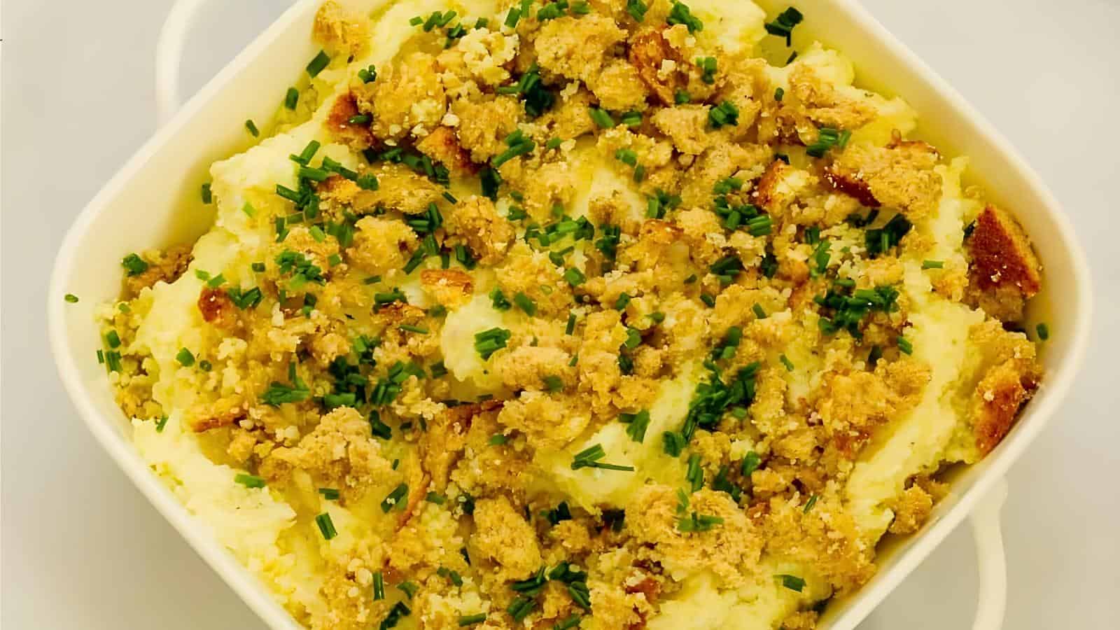 A square white bowl containing mashed potatoes topped with a crunchy breadcrumb mixture and sprinkled with chopped chives.