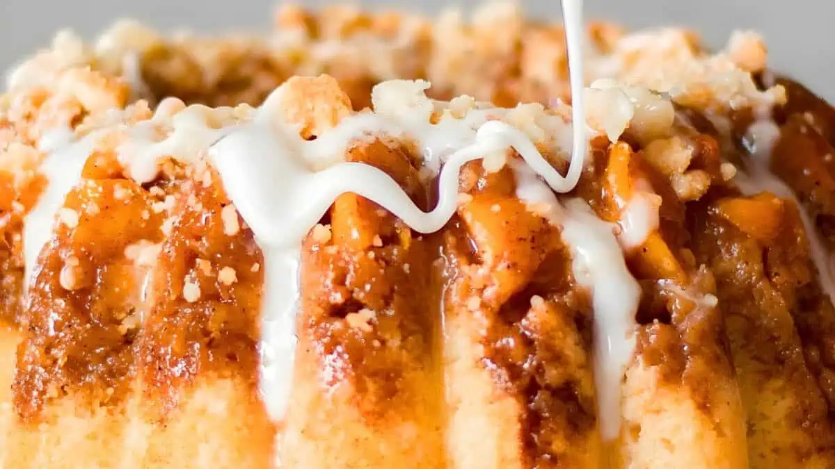 Close-up of a glazed cinnamon apple Bundt cake with visible bits of apple and crumb topping, a ribbon of white icing being drizzled from above. The cake's texture looks moist and inviting with a glossy finish from the glaze.