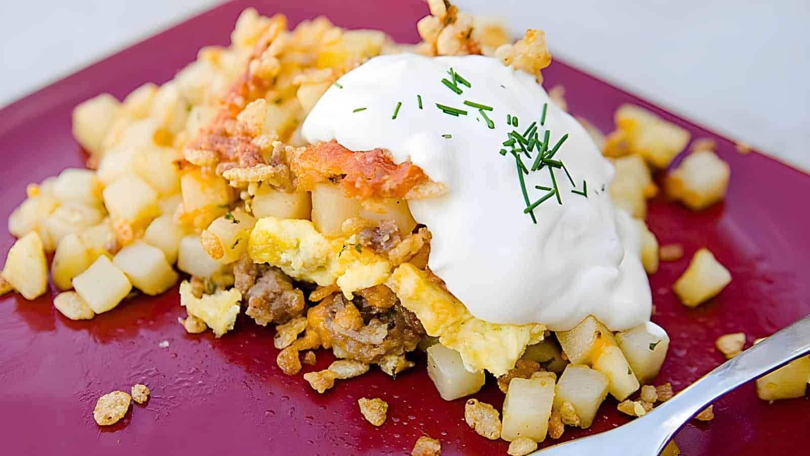 A plate of hash browns topped with scrambled eggs, ground meat, sour cream, and chopped chives next to a fork on a red surface.