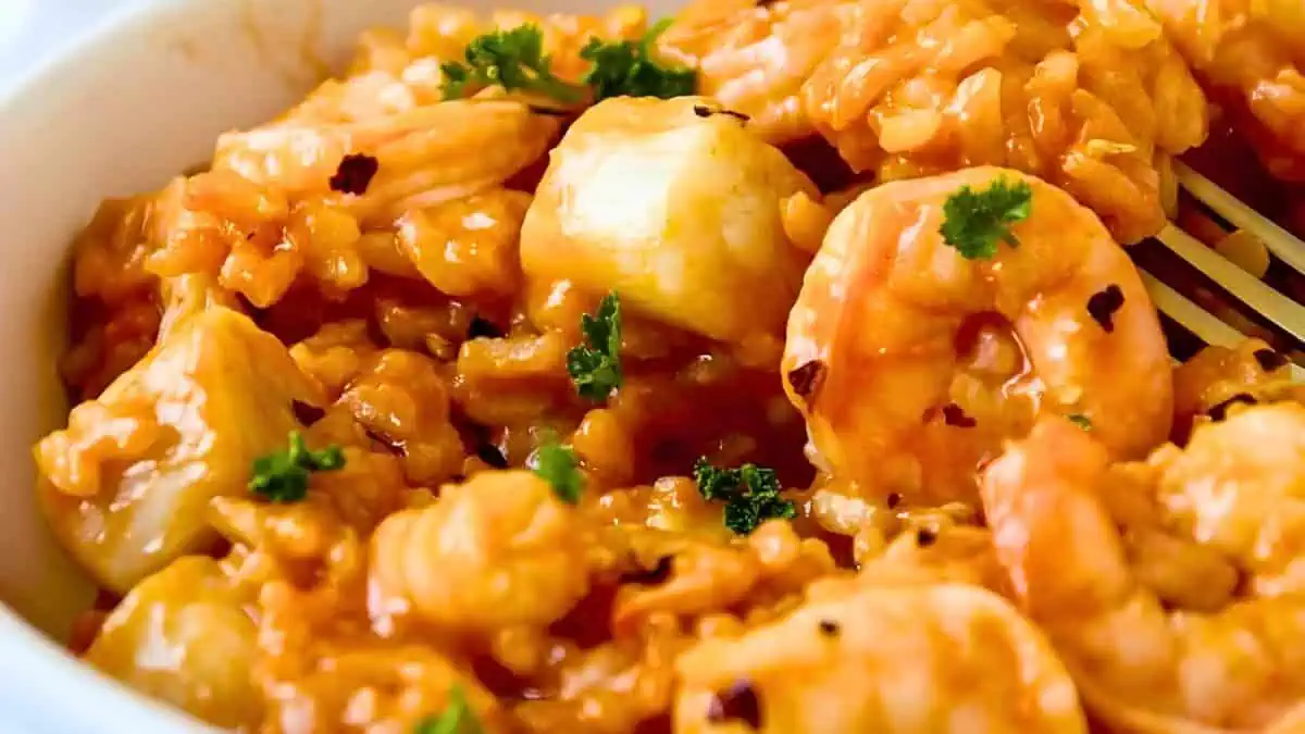 Close-up of a bowl filled with seafood risotto, prominently featuring shrimp and diced scallops mixed with a rich, creamy sauce. Fresh parsley is sprinkled on top as a garnish. A fork is partially visible, ready to dig in.