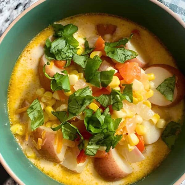 A bowl of chowder with chunks of potato, corn, carrots, and herbs on top, placed on a black and white marble surface next to a green and white checkered cloth.