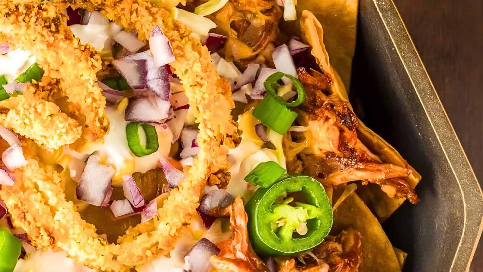 A tray of nachos topped with shredded chicken, melted cheese, diced red onions, green onions, fried onion rings, and jalapeño slices.