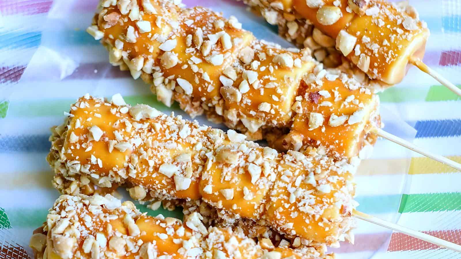 Caramel-covered marshmallow on sticks, coated with chopped peanuts, arranged on a colorful striped surface.