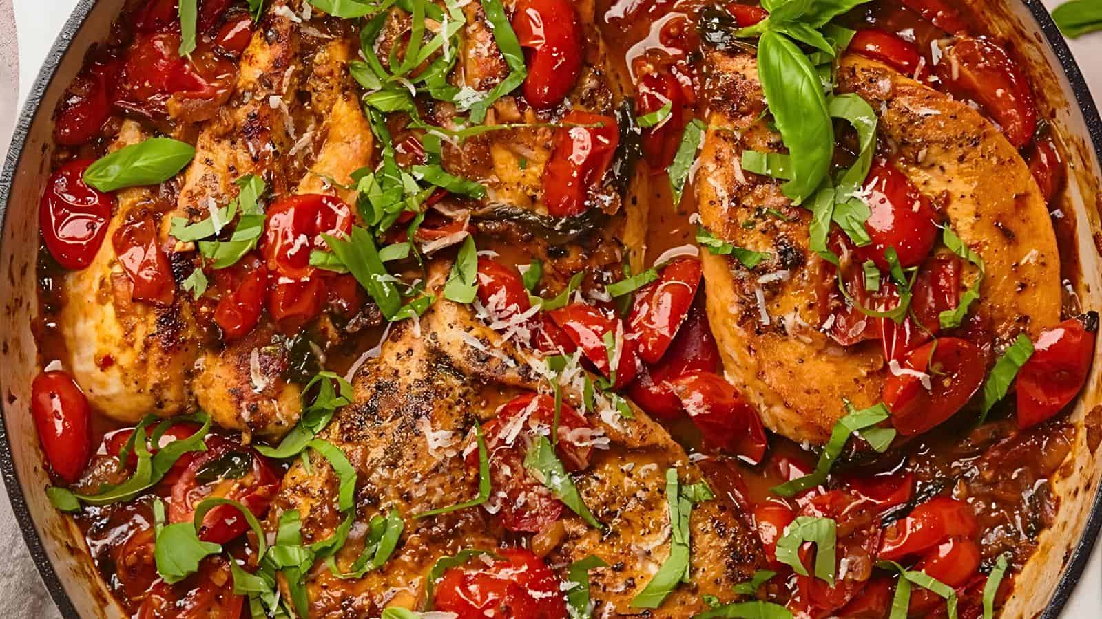 A skillet with cooked chicken breasts, cherry tomatoes, fresh basil, grated cheese, and herbs.