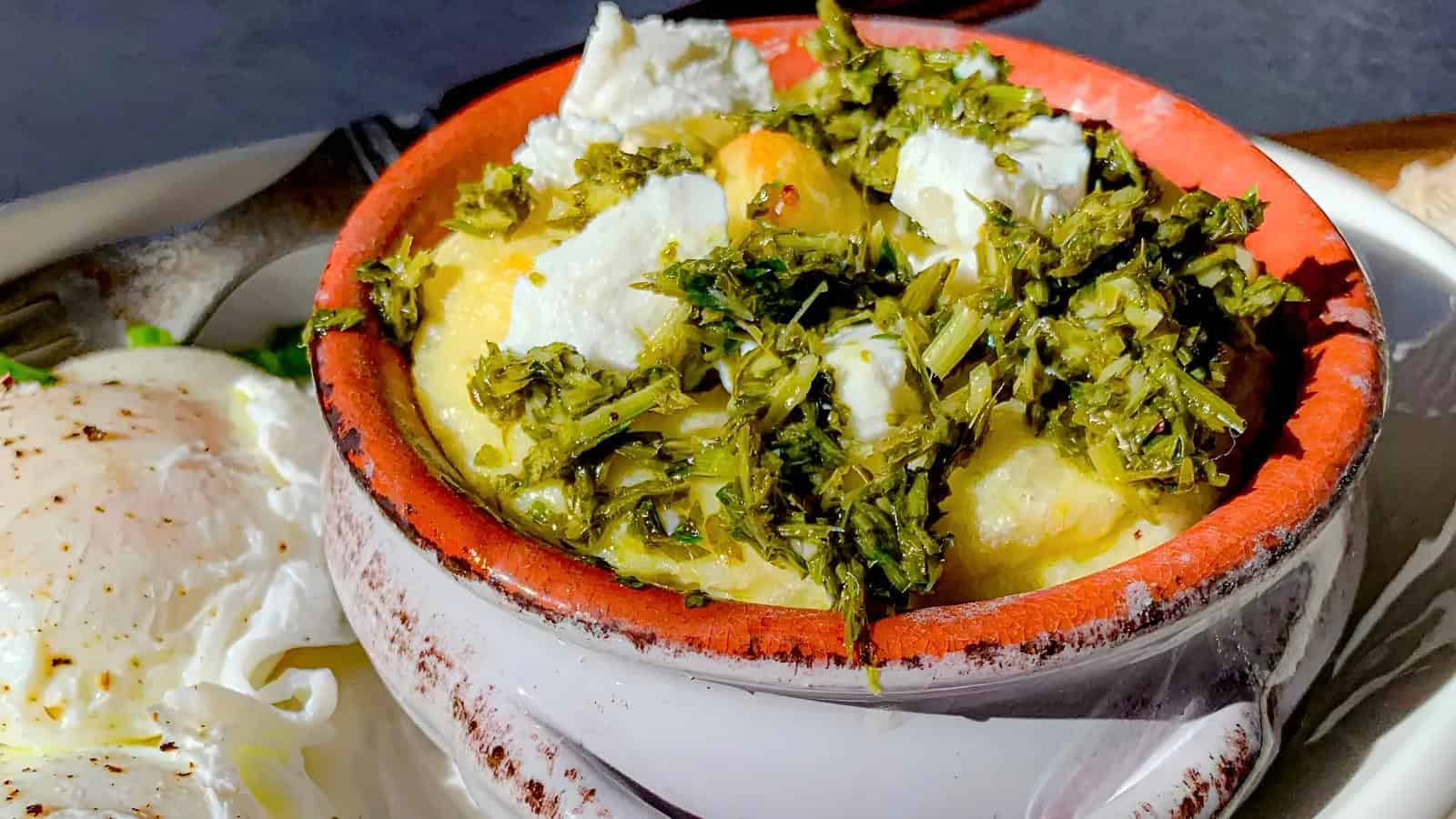 A bowl of mashed potatoes topped with crumbled cheese and chopped green herbs, next to a plate with poached eggs.