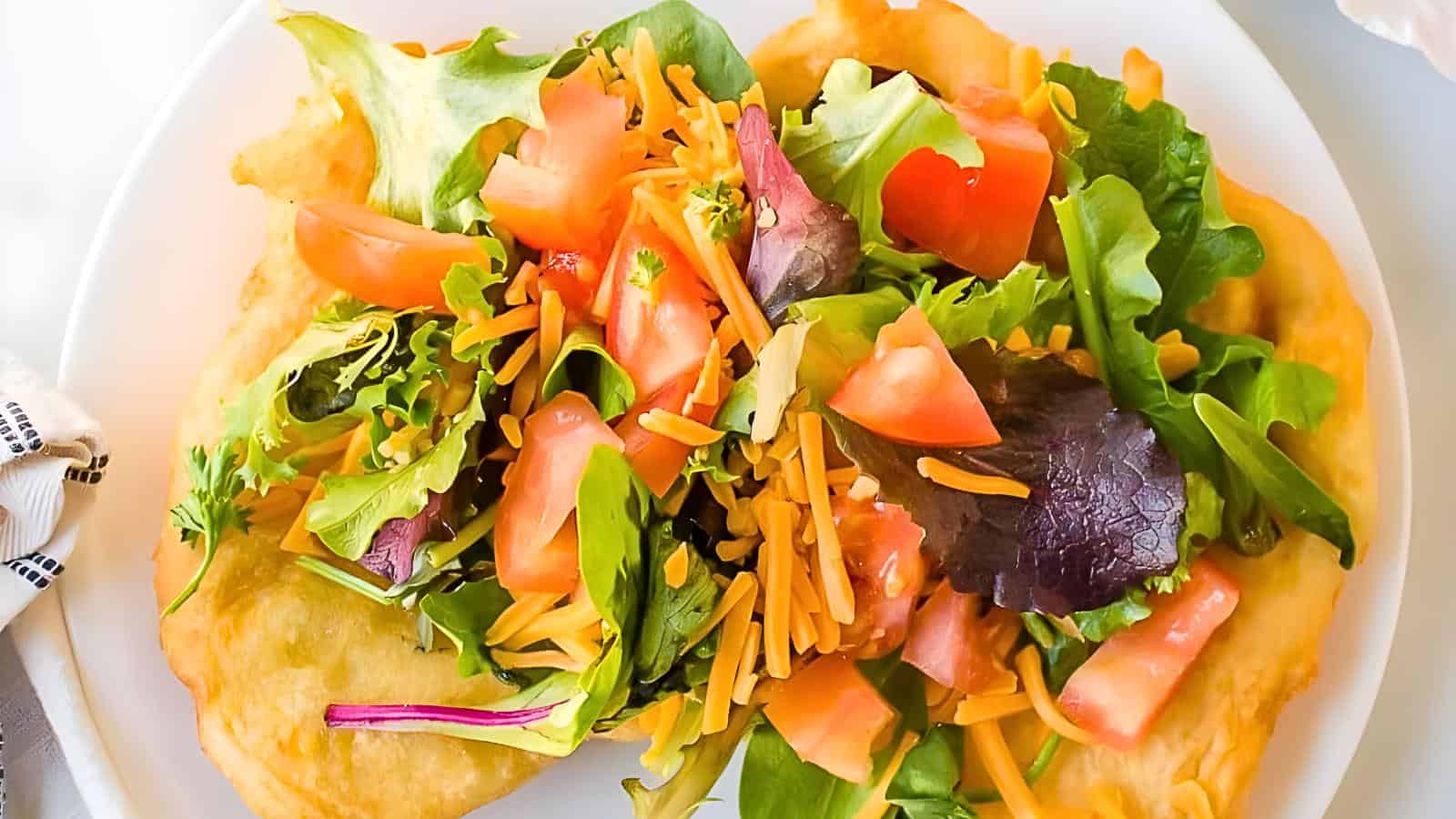 A plate with fry bread, mixed greens, diced tomatoes, and shredded cheese.