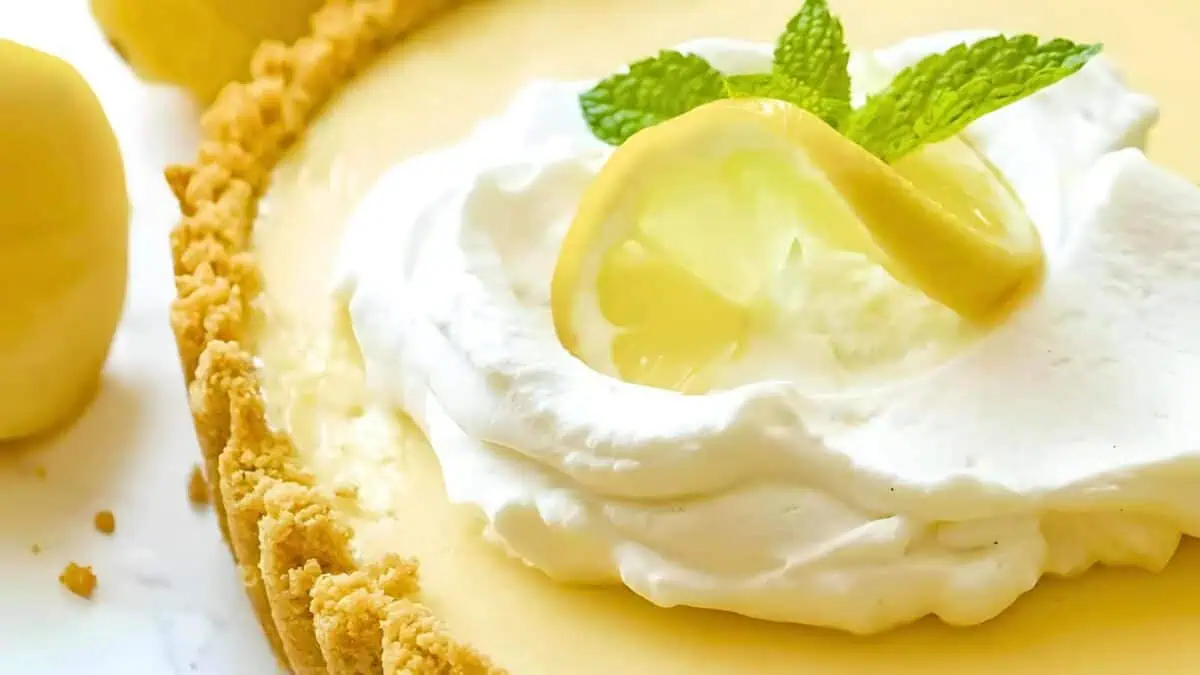 Close-up image of a creamy lemon tart with a golden-brown crust. The tart is topped with a dollop of whipped cream, a slice of lemon, and a sprig of fresh mint. The texture of the crust and the smoothness of the filling are clearly visible.