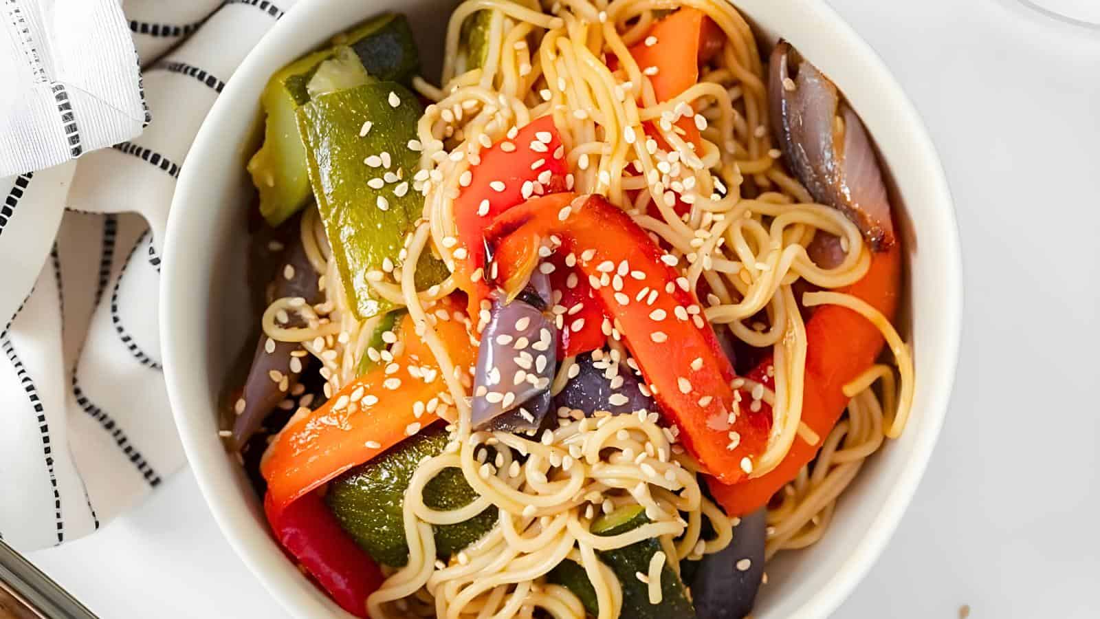 A bowl of stir-fried noodles with colorful bell peppers, zucchini, and red onions, garnished with sesame seeds. A striped cloth napkin is partially visible beside the bowl.