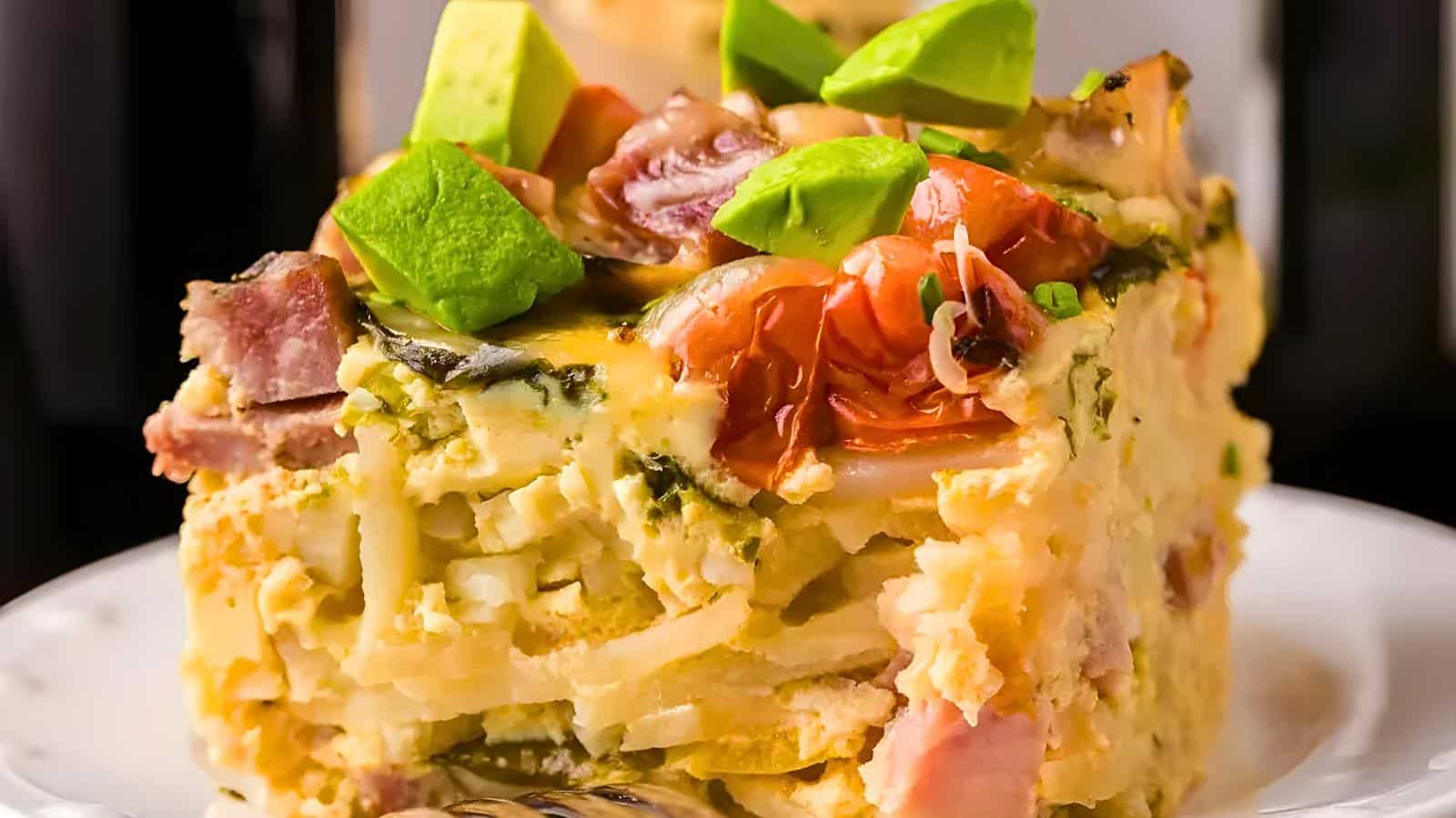 A casserole slice on a plate, topped with diced avocado, cherry tomatoes, and herbs. Visible chunks of meat, vegetables, and cheese are layered in the casserole.