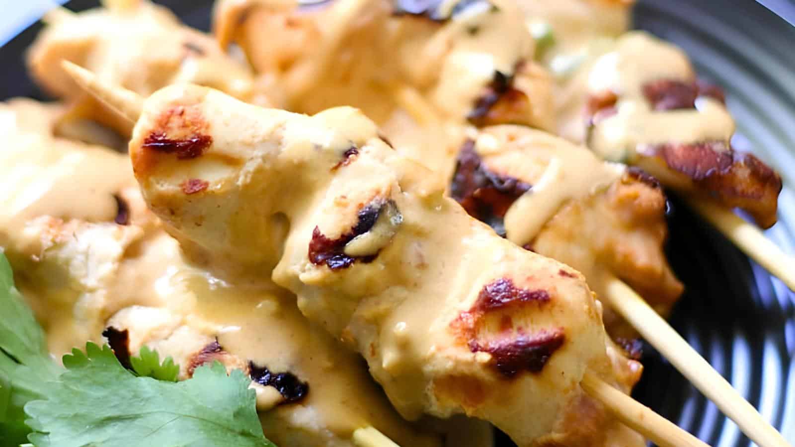 Close-up of grilled chicken skewers drizzled with a creamy sauce, garnished with fresh cilantro leaves on the side. The skewers are served on a black plate, highlighting their golden brown color and charred grill marks.