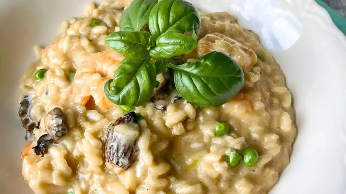 A bowl of creamy risotto garnished with fresh basil leaves. The risotto contains peas, mushrooms, and shrimp, creating a rich and savory dish. The colorful ingredients stand out against the creamy rice base, offering a visually appealing and appetizing meal.
