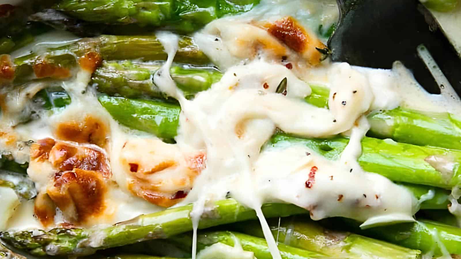 Asparagus spears covered in melted cheese and creamy sauce, with a fork partially visible on the right.