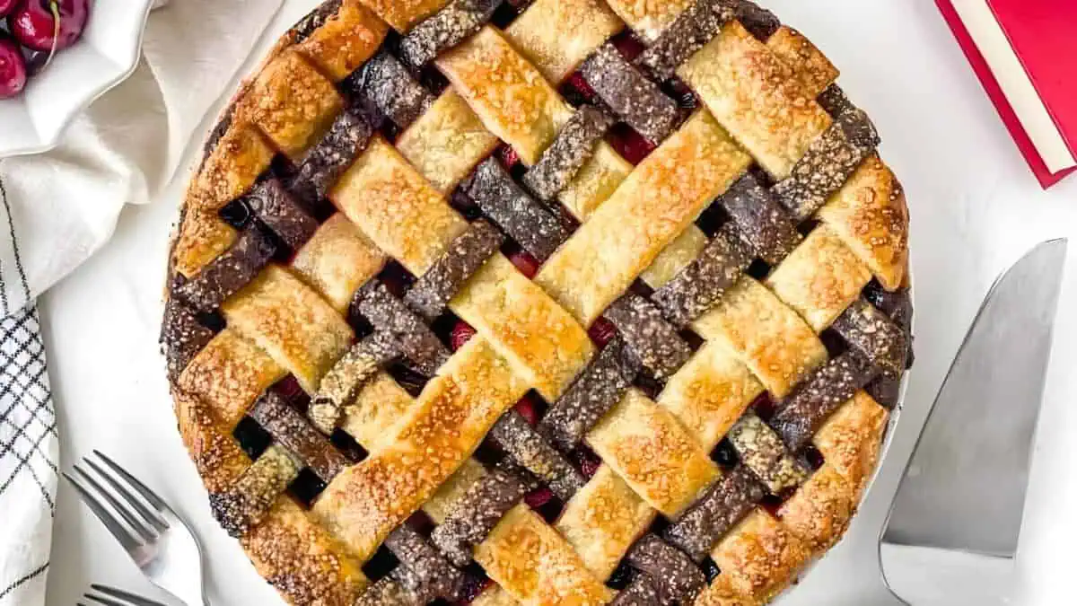A top-down view of a cherry pie with a golden-brown lattice crust. The pie is placed on a white surface, with a pie server, two forks, a checkered napkin, and a few cherries visible nearby. A small red book is partially in view on the right.