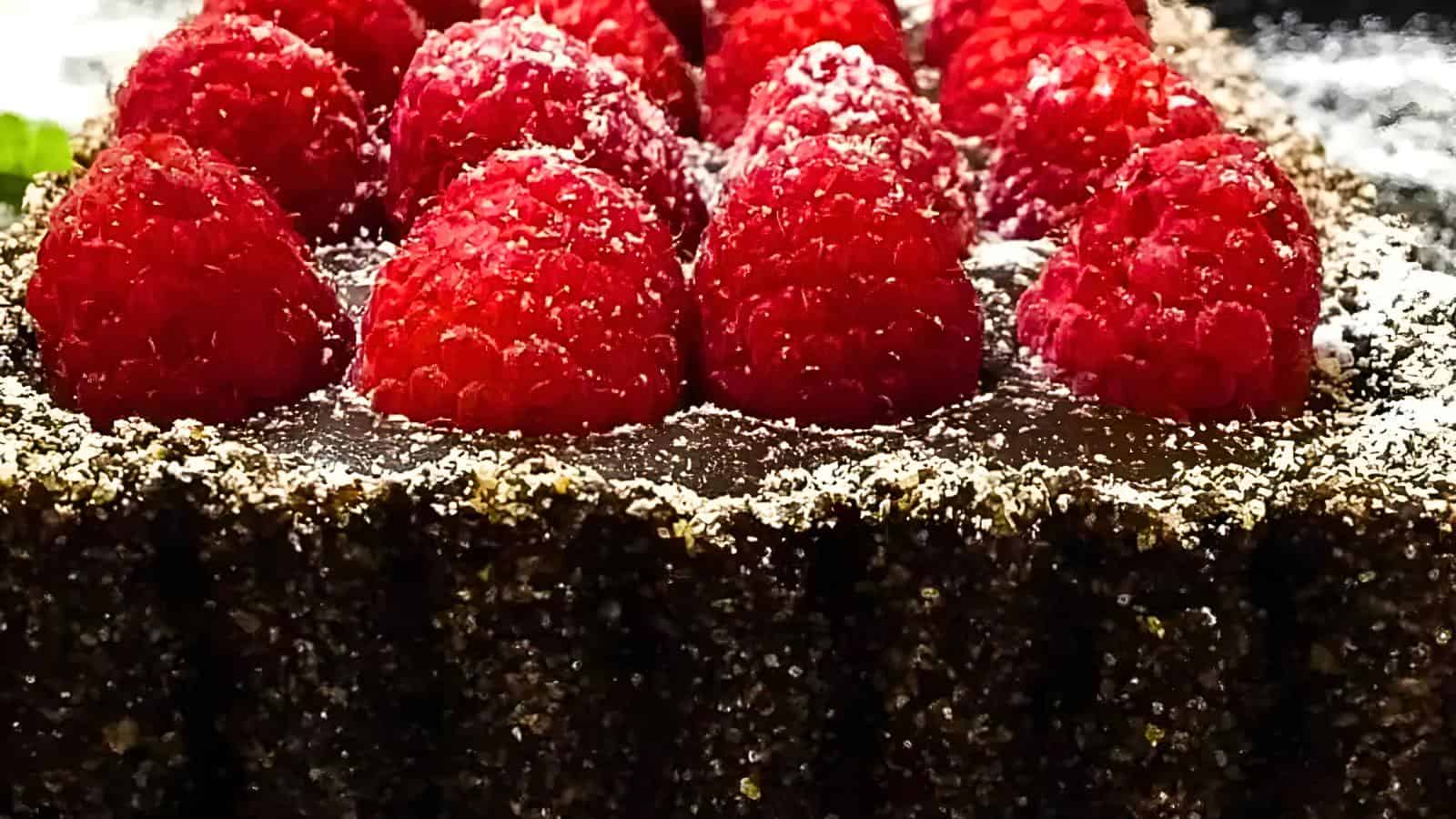 A close-up of a chocolate tart topped with fresh raspberries and a dusting of powdered sugar. The tart has a slightly grainy crust, and the vibrant red raspberries stand out against the rich, dark chocolate filling.
