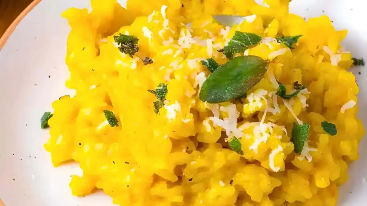 A close-up of a plate of vibrant yellow risotto, garnished with grated cheese and fresh green herbs. The texture of the risotto appears creamy and well-cooked.