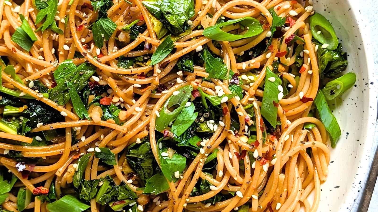A plate of spaghetti mixed with fresh green vegetables, sprinkled with sesame seeds and red pepper flakes, garnished with herbs and drizzled with a light sauce. The dish is vibrant and colorful, served on a white speckled plate.