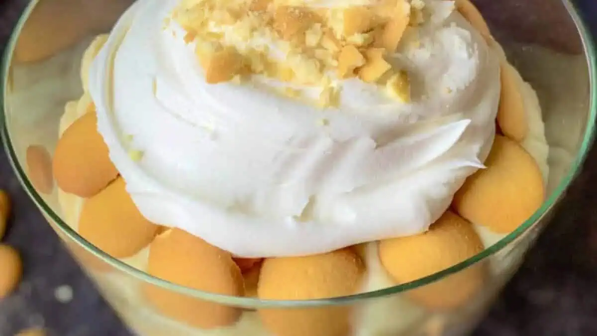 A layered banana pudding dessert is displayed in a glass dish. The top layer consists of whipped cream sprinkled with crushed vanilla wafer cookies, with whole wafers visible around the edges. The dessert appears creamy and inviting.