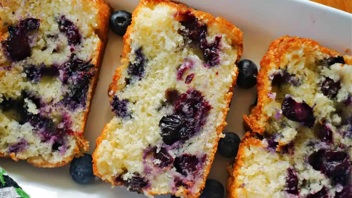 Close-up of three slices of blueberry bread on a white rectangular plate. The bread is golden brown at the edges with a light, moist interior filled with numerous blueberries. Some scattered blueberries surround the slices on the plate.