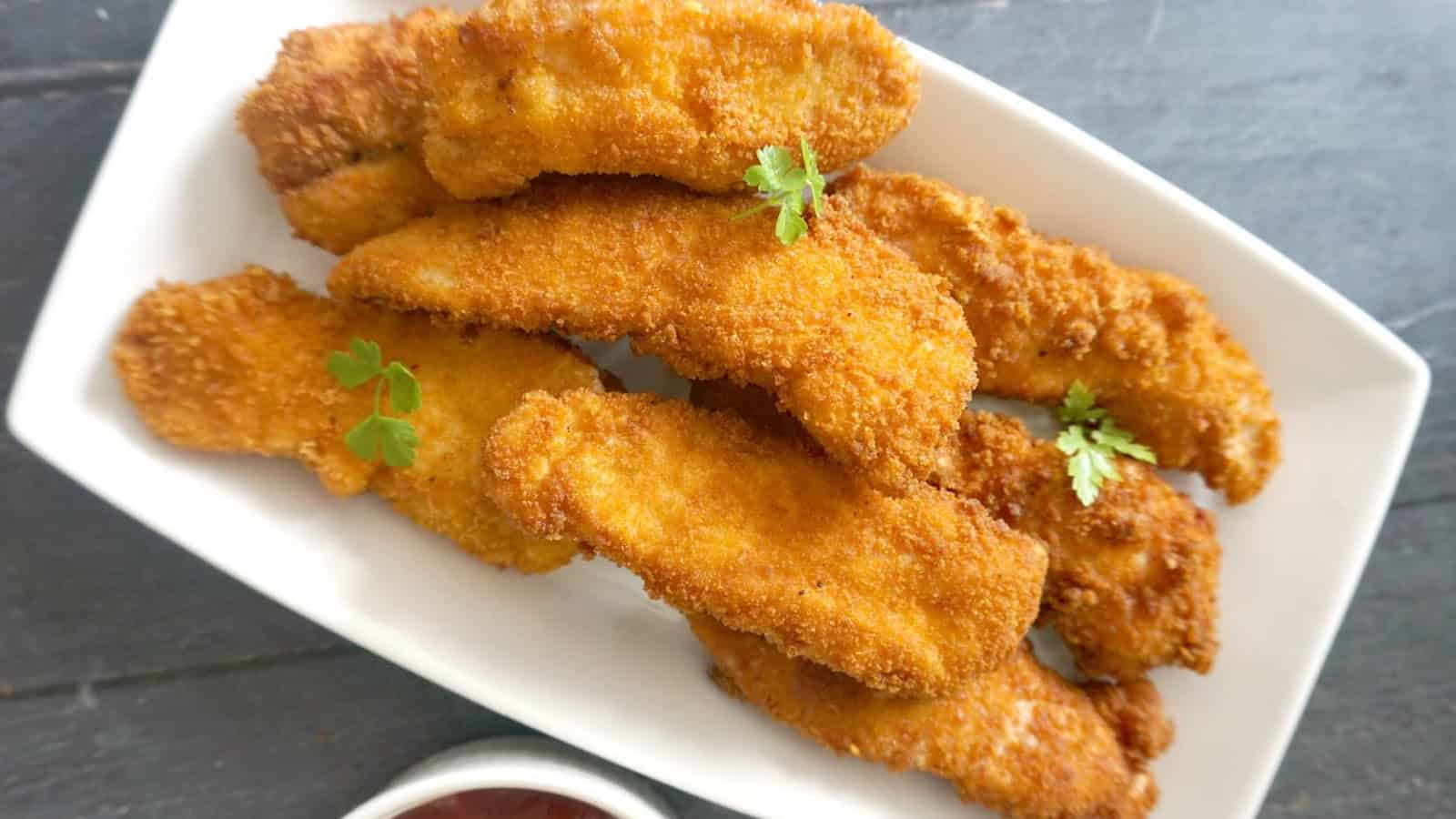 A white rectangular plate holding several breaded and fried chicken tenders, garnished with a few sprigs of parsley.