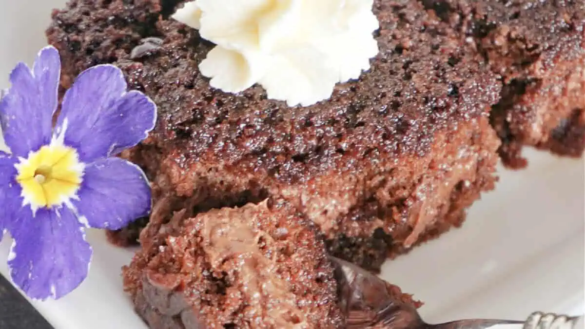 A close-up of a piece of chocolate lava cake on a white plate. The cake has a gooey chocolate center, a dollop of whipped cream on top, and is garnished with a purple flower. A fork has taken a bite from the piece, revealing the rich, moist inside.