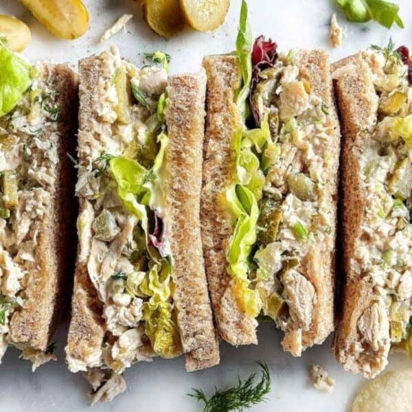 Four chicken salad sandwiches with lettuce on whole wheat bread are arranged side by side on a white surface, surrounded by pickles and potato chips.