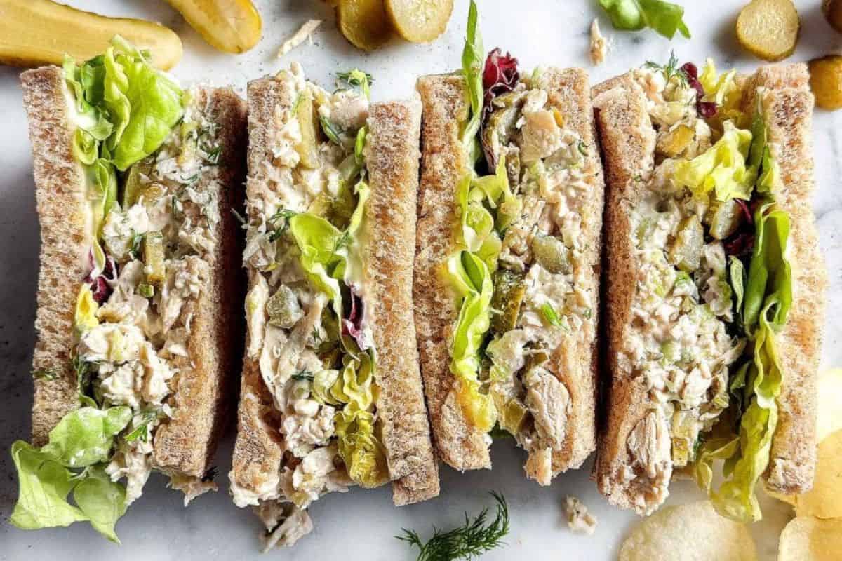Four chicken salad sandwiches with lettuce on whole wheat bread are arranged side by side on a white surface, surrounded by pickles and potato chips.