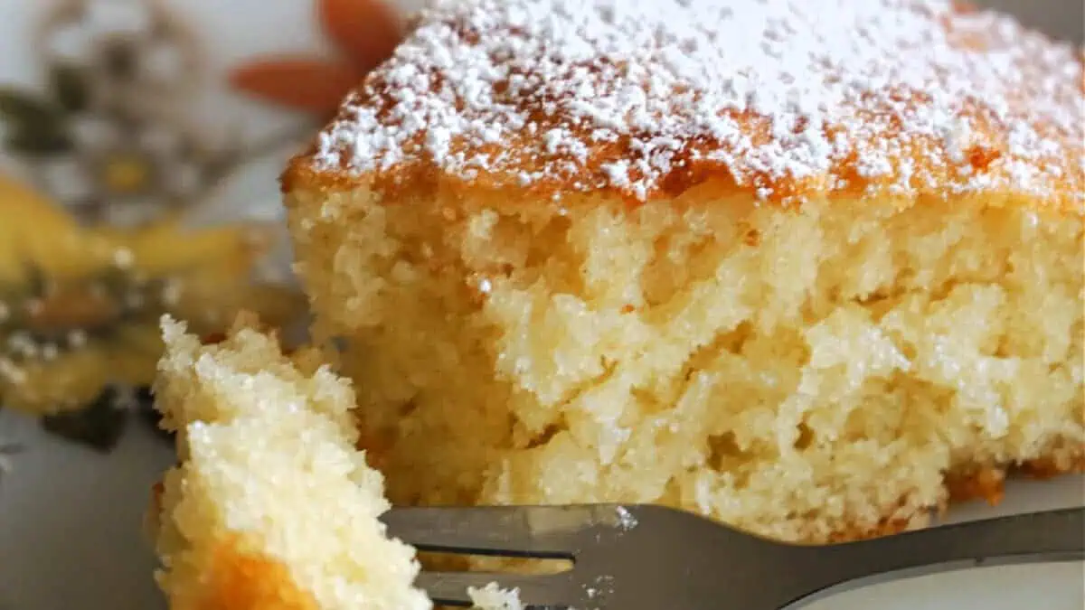A close-up of a slice of fluffy vanilla cake dusted with powdered sugar. A fork has taken a bite out of the cake, revealing its soft and moist texture. The background shows a patterned plate partially visible.
