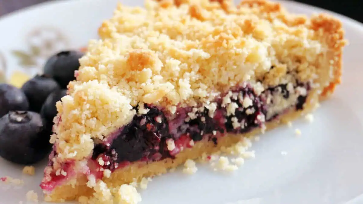 A slice of blueberry crumble pie on a white plate. The pie features a golden, crumbly topping and a rich, moist blueberry filling. Blueberries are garnished beside the slice.