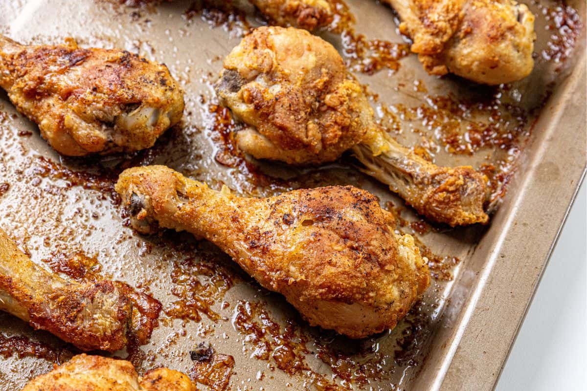 Several baked chicken drumsticks with browned, crispy edges are arranged on a metal baking sheet.
